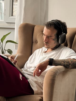 Man wearing headphones seated in a tan chair displays a large dark tattoo covering his right arm and shoulder.