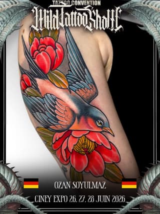 Colorful bird tattoo with red flowers on upper arm, featuring orange and blue wing details with yellow accents and a yellow eye.