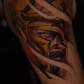 Detailed portrait tattoo of a bearded man wearing a golden helmet with dark shading, displayed on an arm with warm brown and gold tones.