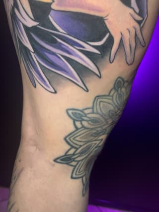 Colorful tattoo on an arm featuring purple and blue feathered wings at the top and a geometric mandala-inspired flower design below in teal.
