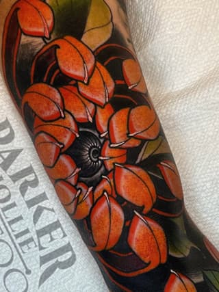 Red and orange flower with black center and dark background on forearm, featuring detailed petals with shading and green leafy elements.