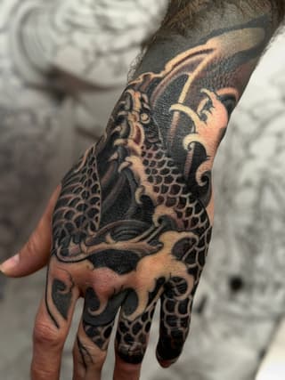 Dragon tattoo covering the back of a hand and forearm in black and grey ink with detailed scales and ornamental swirls.