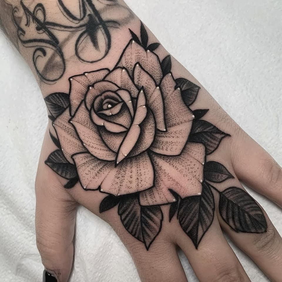 Large rose with detailed petals and eye detail in center, surrounded by leaves, tattooed on hand with dotted shading and black linework.