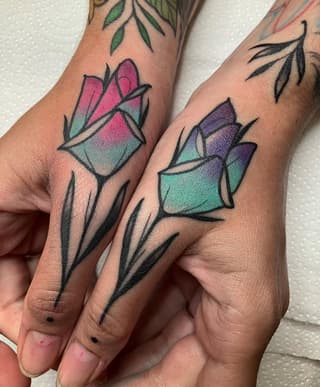 Two hands displaying matching flower tattoos on the knuckles with pink, purple, and teal watercolor fills and black outlines.