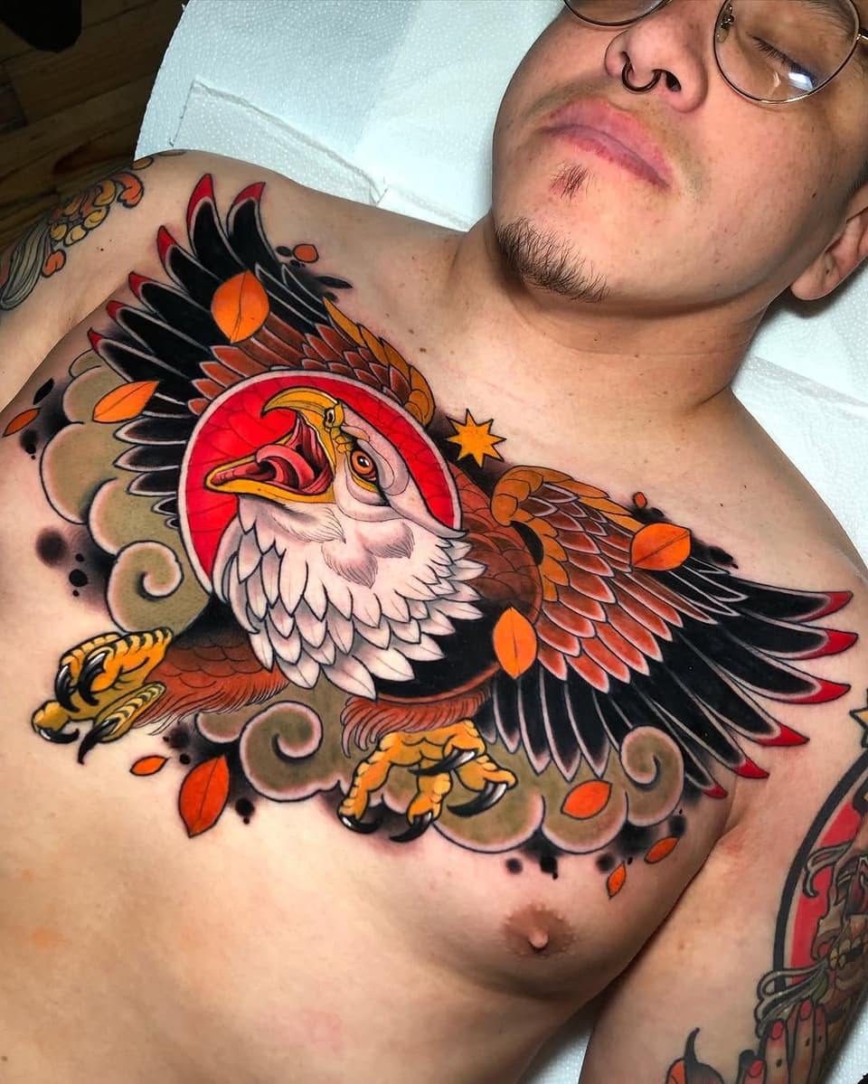 Colorful eagle chest tattoo featuring a red circular background, orange and black wings, white chest feathers, decorative scrollwork, and falling leaves in orange and red tones.