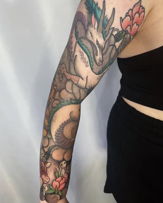 Full-color arm sleeve tattoo featuring anatomical heart with flowers, botanical elements, and small skeletal figures in coral, green, and tan tones.