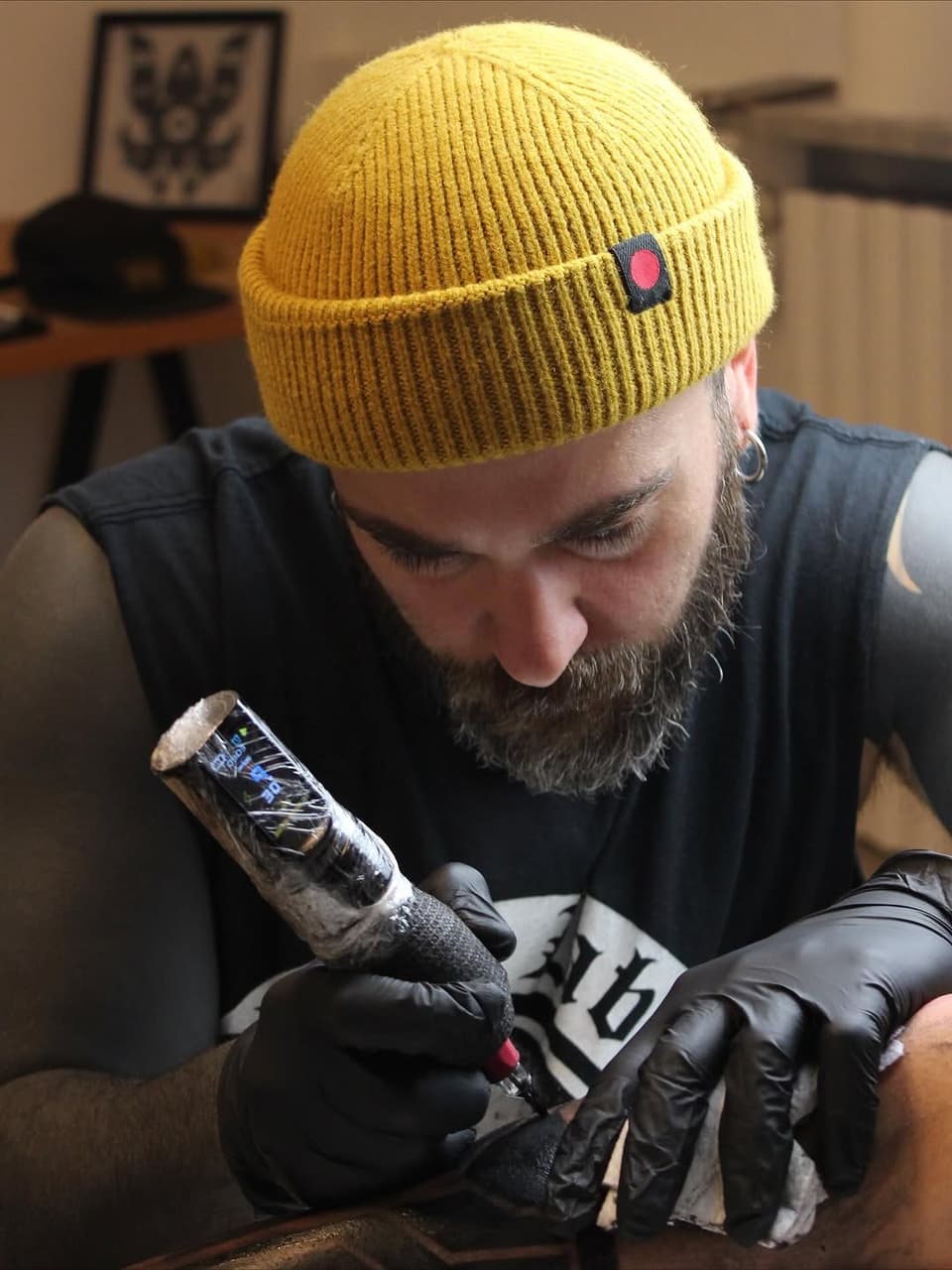 Bearded tattoo artist wearing a yellow beanie and black gloves examines a fresh tattoo on a client's arm featuring pink and blue colors.