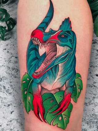 Colorful dinosaur head tattoo with open jaws in teal and red, surrounded by green tropical leaves on the upper leg.