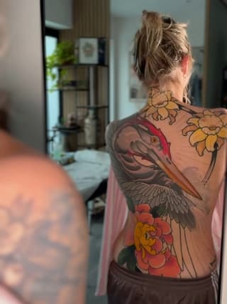 Woman displaying large back tattoo featuring colorful flowers, a bird with wings, and a rose in yellow, red, orange, and grey tones.