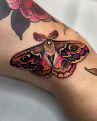 Colorful moth tattoo with burgundy, orange, and gold wings featuring eye-like markings, positioned on the upper arm.