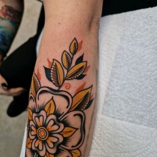 Colorful floral tattoo on forearm featuring yellow and orange flowers with black leaves and ornamental swirls against skin.