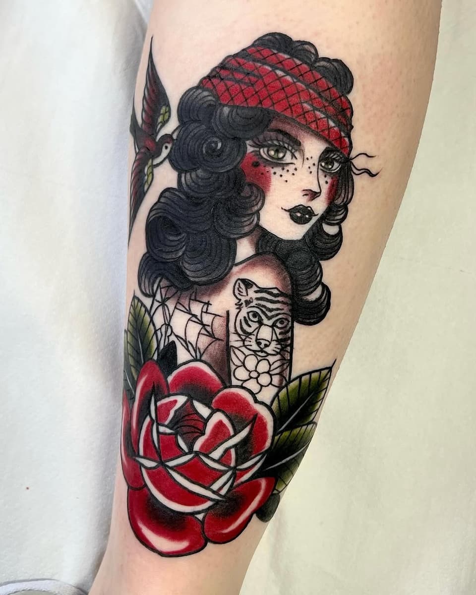 Woman's portrait with dark curled hair and red headwrap wearing tiger tattoo on shoulder, surrounded by red rose below, on upper arm.