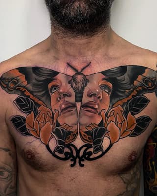 Symmetrical chest tattoo featuring mirrored female faces with moth wings in orange and black ink, with ornamental flourishes below.