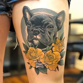 Black French Bulldog head with yellow roses and green leaves on thigh, with "BRUCE" text on a bone charm below the flowers.