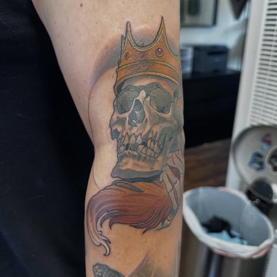 Skull wearing a golden crown with red hair flowing below, tattooed on an arm in color with orange, gray, and red tones.