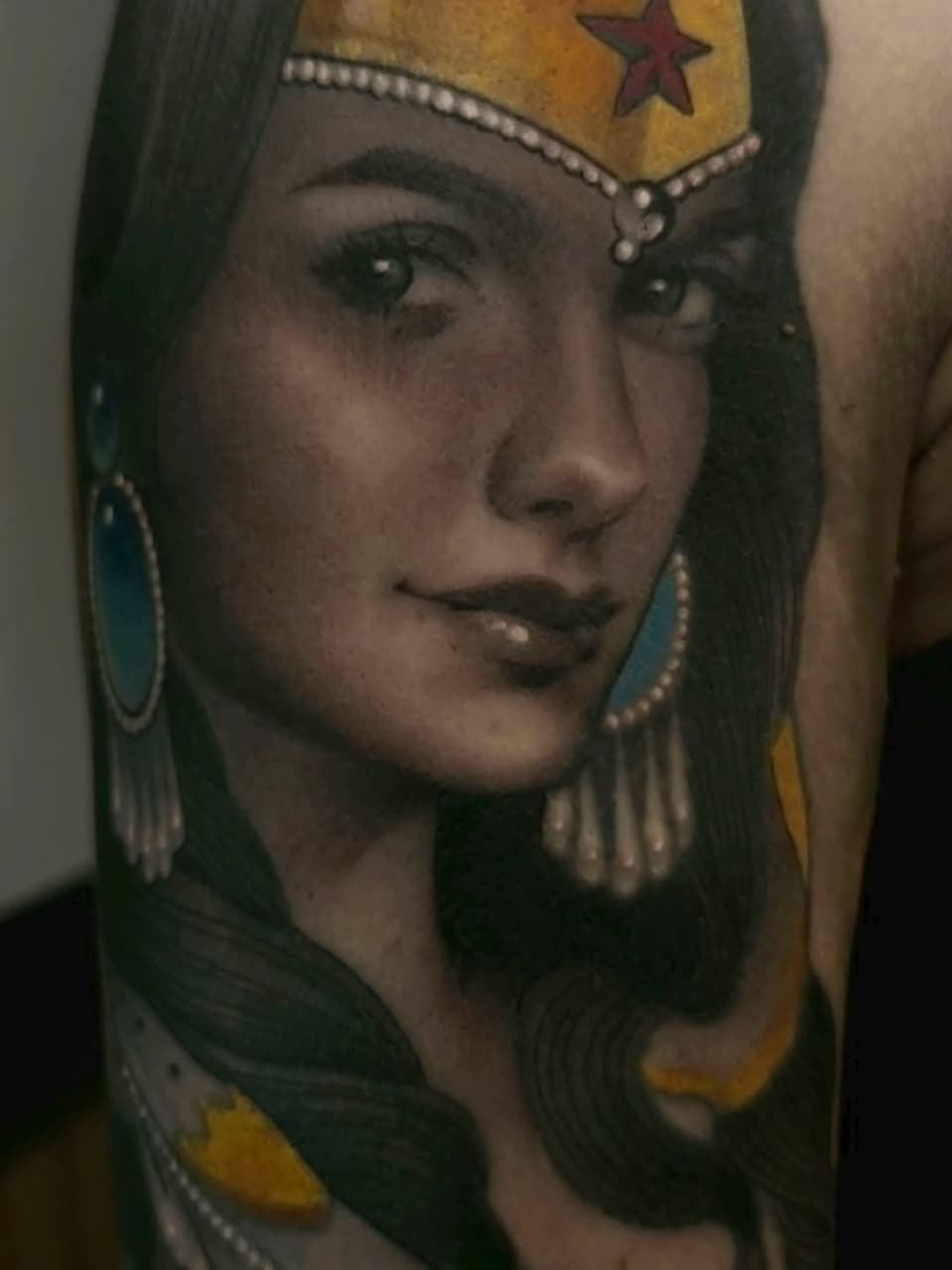 Portrait tattoo of a woman with long dark hair, pearl earrings, and a gold headdress with a red star, on upper arm in color.