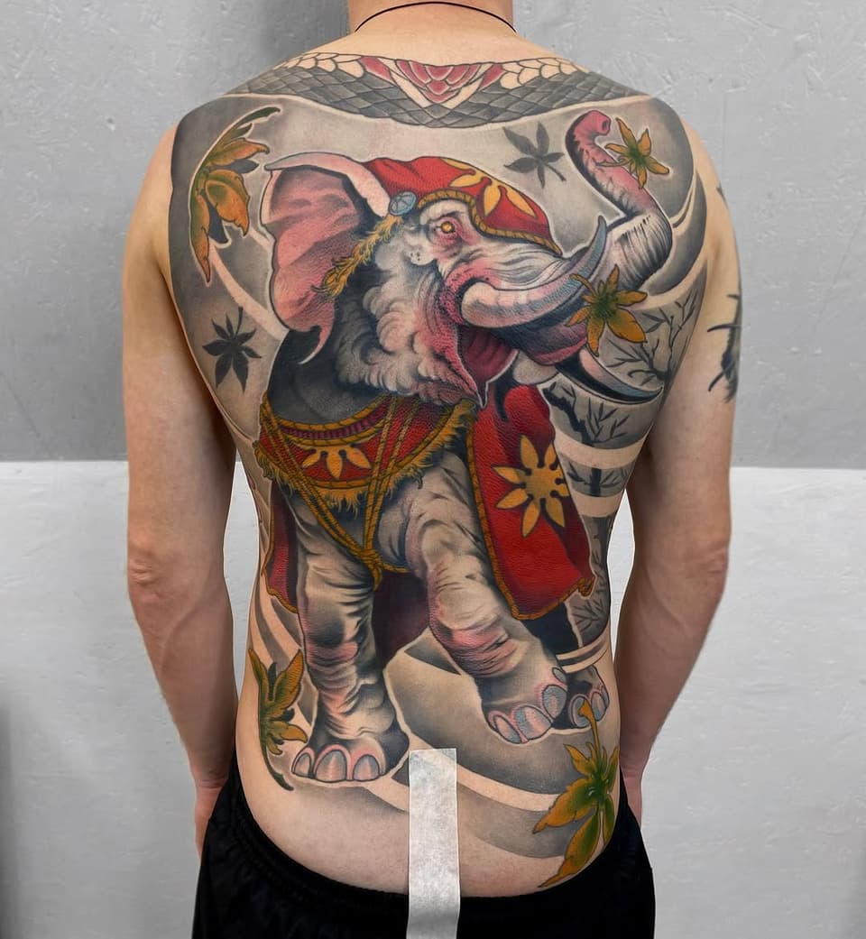 Full-back tattoo of an ornately dressed elephant in red and gold clothing with floral accents, displayed on a shirtless person from behind.