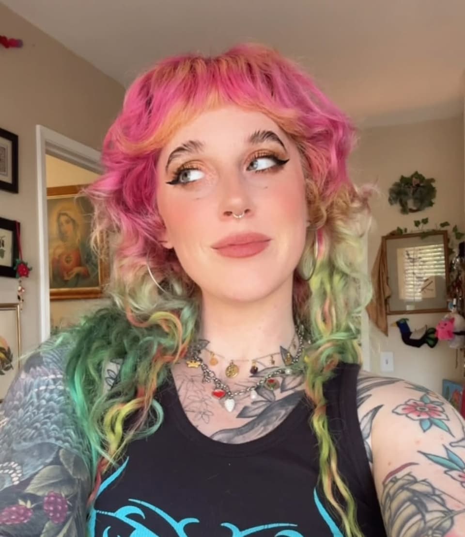 Portrait of a person displaying colorful floral and botanical tattoos on their arms and chest, wearing a black graphic t-shirt.