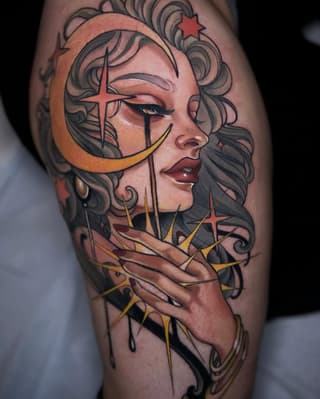 Portrait tattoo of a woman's profile with curled hair, celestial symbols including a crescent moon and crosses, rendered in orange, yellow, and black on the upper leg.