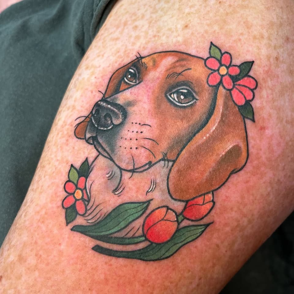 Brown dog's head with expressive eyes adorned with red flowers and green leaves, tattooed on upper arm in red, orange, brown, and green inks.