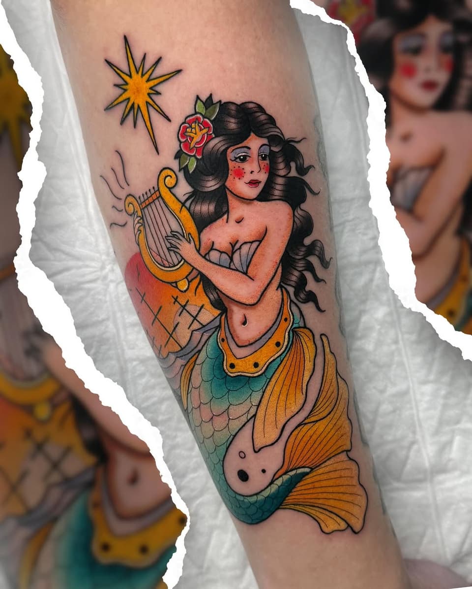 Mermaid holding a golden lyre with a red flower in her hair and a star above, tattooed on the upper arm in orange, teal, and black colors.