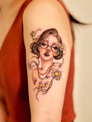 Portrait tattoo of a woman wearing glasses with wavy hair adorned with daisies and decorative vine elements in brown, yellow, and skin tones on the upper arm.