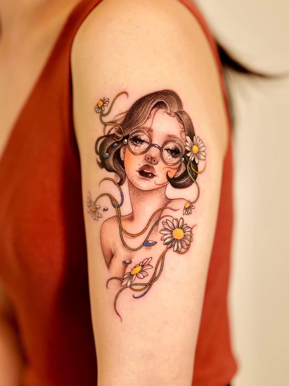 Portrait tattoo of a woman wearing glasses with wavy hair adorned with daisies and decorative vine elements in brown, yellow, and skin tones on the upper arm.