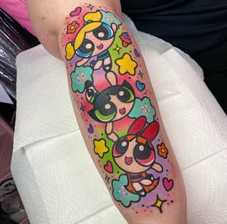 Three cartoon superhero girls with black masks and blonde/dark hair surrounded by colorful stars, hearts, and flowers on a calf tattoo in bright pink, yellow, green, and blue.