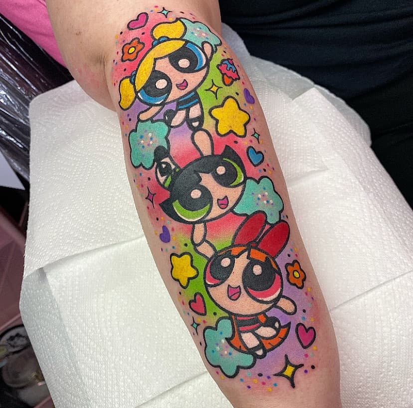 Three cartoon superhero girls with black masks and blonde/dark hair surrounded by colorful stars, hearts, and flowers on a calf tattoo in bright pink, yellow, green, and blue.