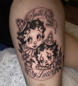 Betty Boop character tattoo with "Smile Now Cry Later" banners on upper arm, rendered in black and gray ink.
