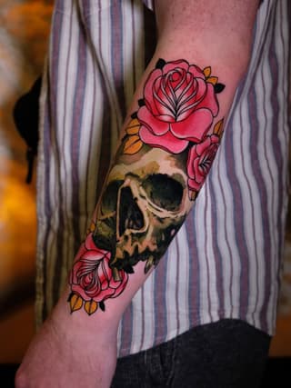Colorful skull tattoo with pink roses and orange leaves on the forearm of a person wearing a striped shirt.