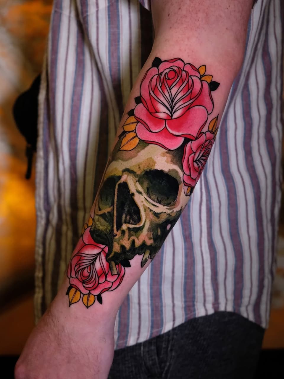 Colorful skull tattoo with pink roses and orange leaves on the forearm of a person wearing a striped shirt.