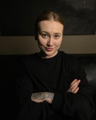 Portrait of a person wearing a black long-sleeved shirt displaying a large ornamental wing tattoo on their forearm in black ink.