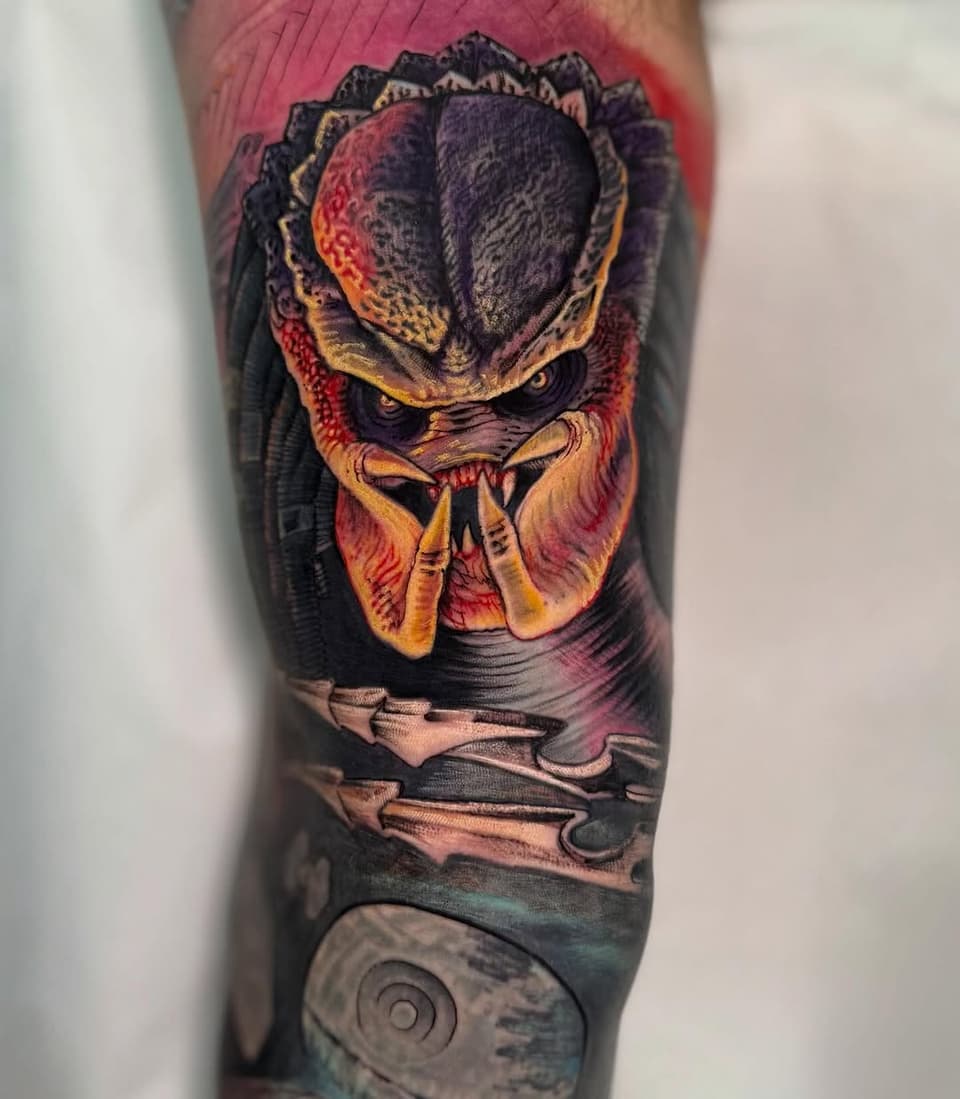 Colorful Predator creature head tattoo on leg featuring red, gold, and black tones with textured details and spiral design at bottom.