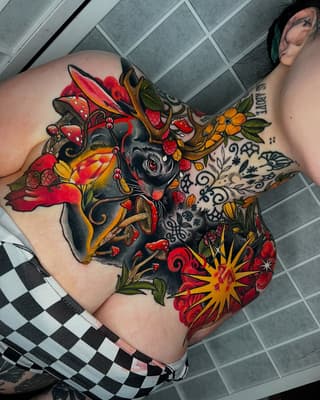 Colorful shoulder tattoo featuring a black cat surrounded by red roses, yellow flowers, mushrooms, and ornamental swirls in red, yellow, orange, and green.