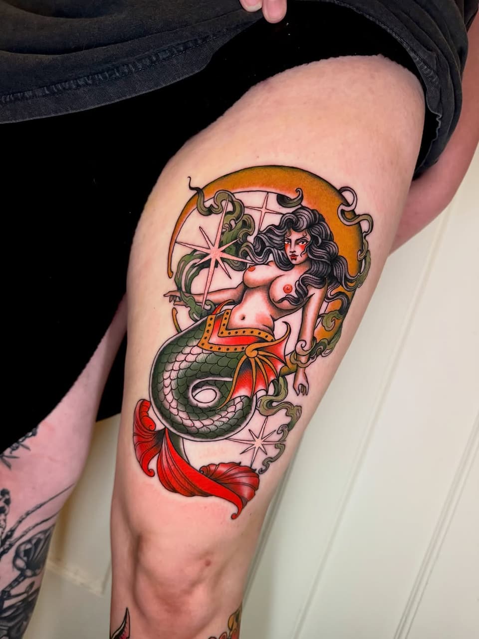 Colorful mermaid tattoo on thigh featuring a woman with dark hair and serpent tail in orange, green, and red with decorative flourishes.