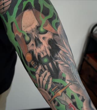 Detailed skull tattoo on arm with green flame designs, orange accents, and black shading on skin.