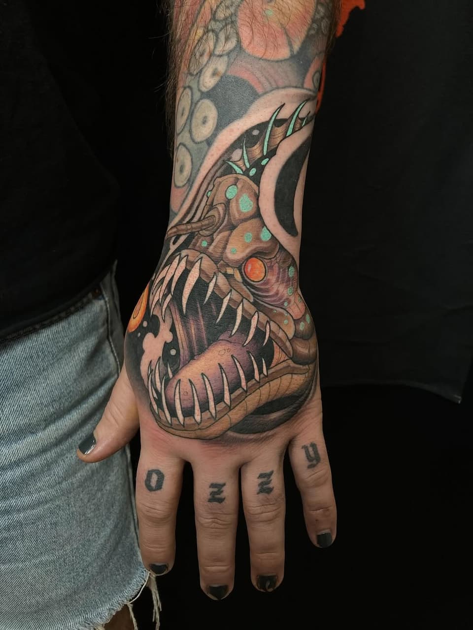 Colorful demon or creature head tattoo on hand and forearm with sharp teeth, horns, and ornamental details in orange, green, and burgundy tones.