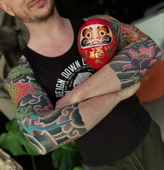 Man displaying full-sleeve arm tattoos featuring colorful flowers, dragons, and ornamental designs, holding a red Daruma doll with gold characters.