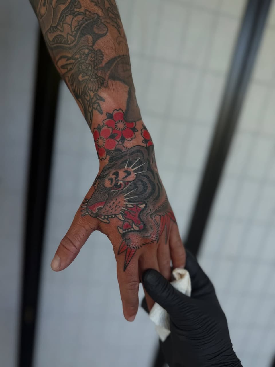 Tattooed hand and arm featuring a tiger face on the hand, red flowers on the forearm, and dragon designs on the upper arm in black and red ink.