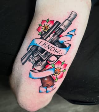 Revolver with wooden grip and blue banner reading "I KNOW" surrounded by red flowers on calf muscle.