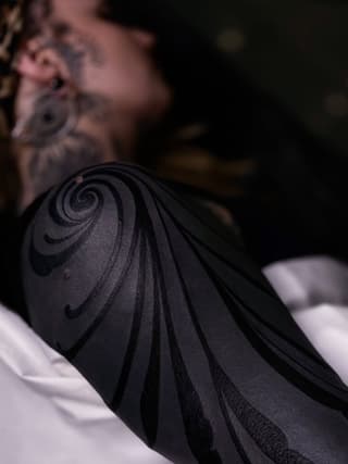 Spiral and curved line tattoo in black ink on a person's shoulder, with blurred background showing additional body art.