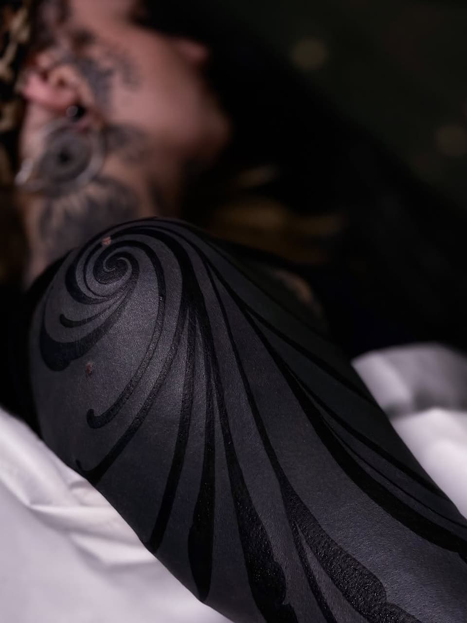 Spiral and curved line tattoo in black ink on a person's shoulder, with blurred background showing additional body art.