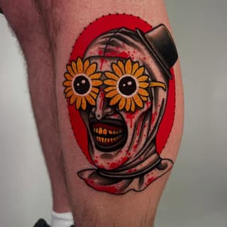 Colorful skull tattoo on a calf with sunflower eyes, top hat, open mouth showing golden teeth, red background, and ribbon banner details.