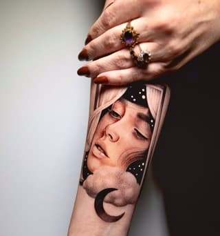 Portrait tattoo on forearm featuring a contemplative face with closed eyes, crescent moon, stars, and decorative elements in brown and black ink.