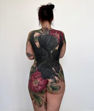 Back tattoo featuring a black raven with spread wings surrounded by large red peonies and green foliage in full color.