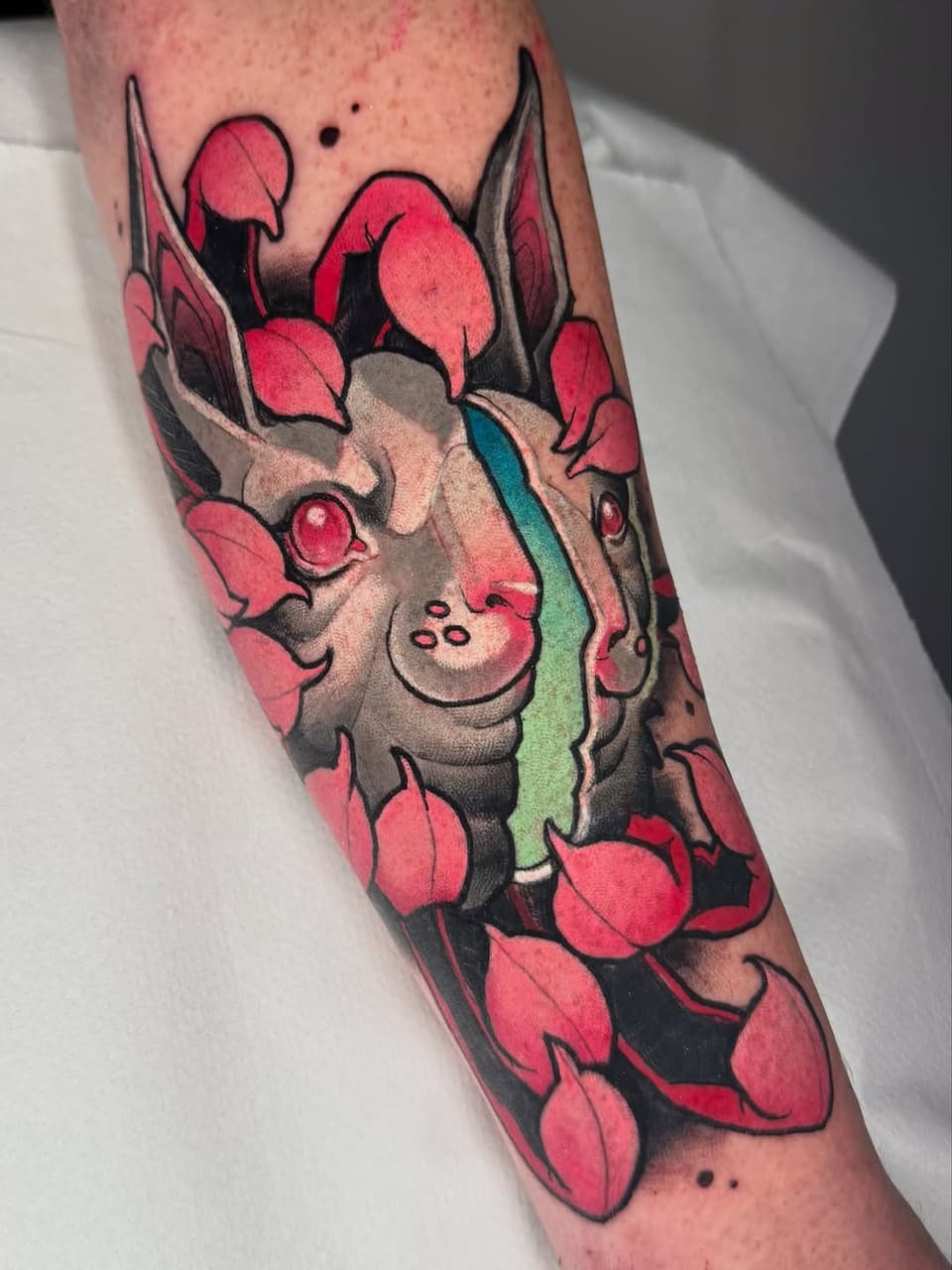 Colorful tattoo on forearm featuring a horned creature surrounded by red petals and flowers with green and tan shading details.