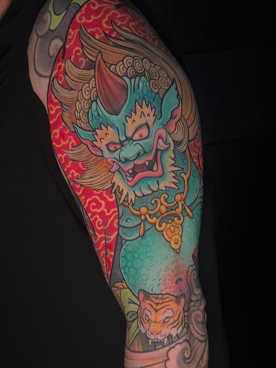 Colorful full-sleeve tattoo featuring a turquoise demon mask with red accents, golden ornamental details, winged elements, and a tiger head lower on the arm.