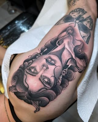 Detailed portrait tattoo on upper arm featuring a face with closed eyes, ornate jewelry, and flowing hair in black and pink shading.