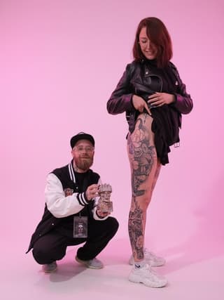 Tattoo artist displays extensive leg piece featuring ornate designs and decorative elements in dark ink on client against pink background.